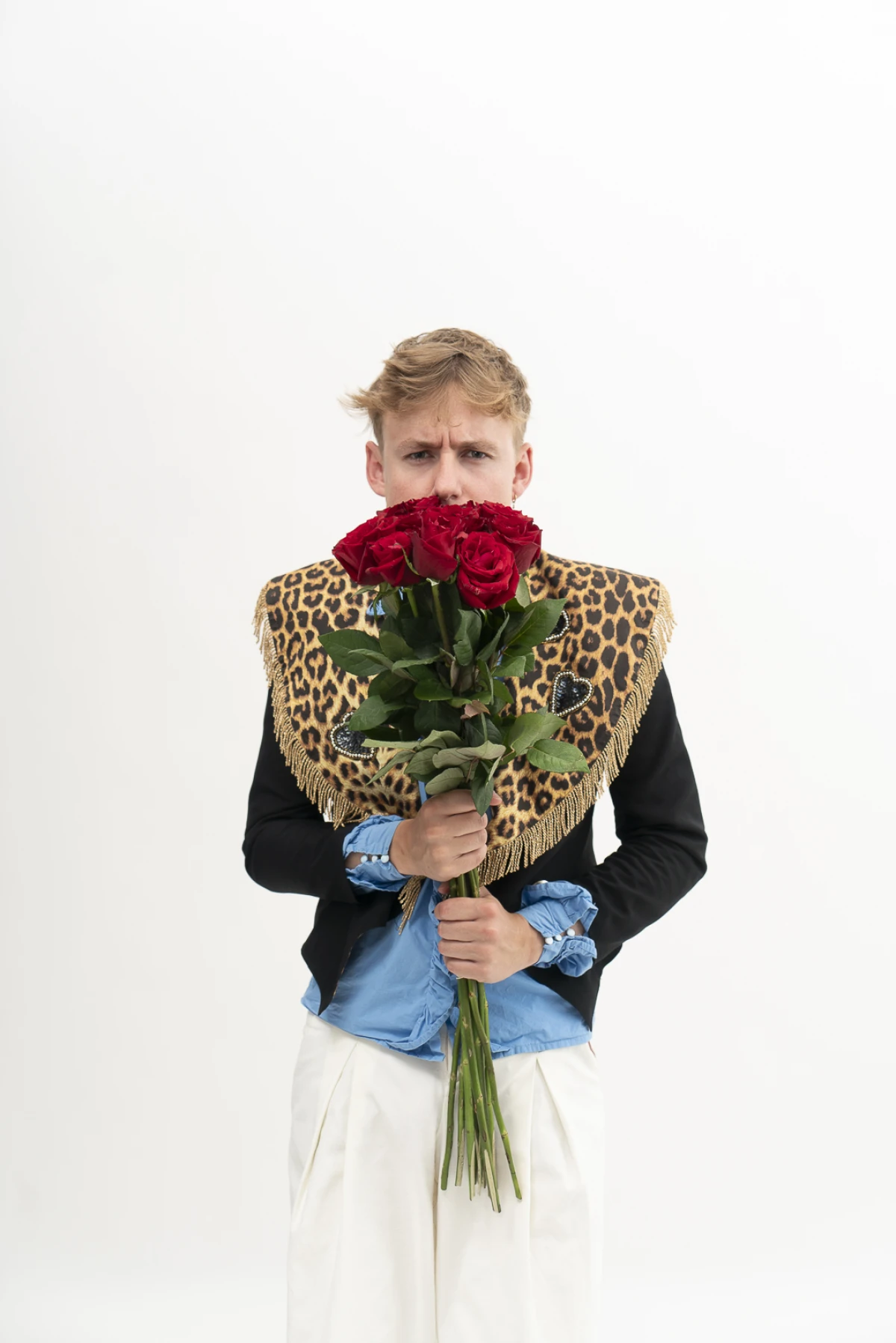 A young man wearing a leopard-print fringed shawl holding a bouquet of red roses over the lower half of his face against a plain white background.